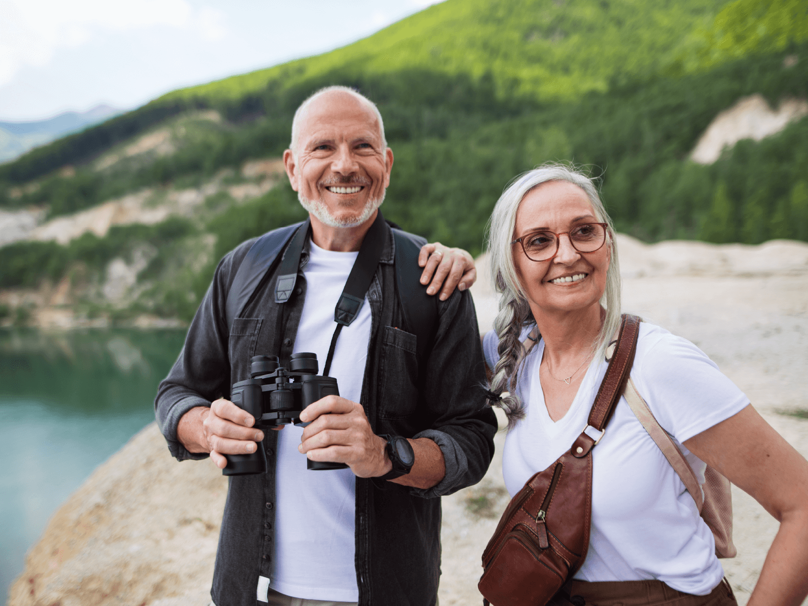 An older white man and woman are smiling while on a hike. The man is holding a pair of binoculars and has a beard. The woman has a sling bag and wears glasses and has her hair in a braid. The background is a mountain with green trees and a lake below. 