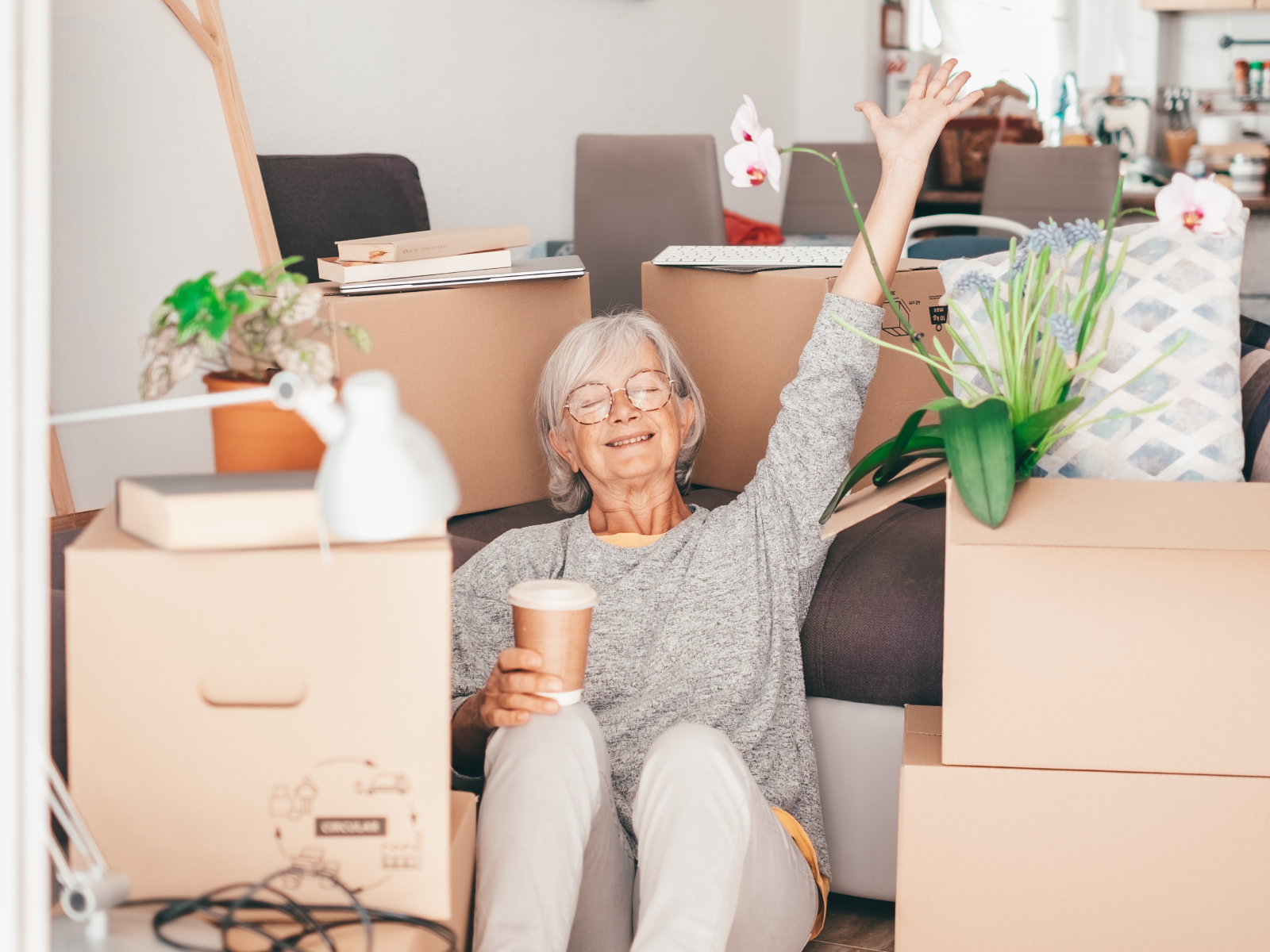 An older woman with grey hair and glasses is wearing a grey shirt and pants. She's sitting surrounded by moving boxes, with one hand in the air and another hand holding a cup of coffee.